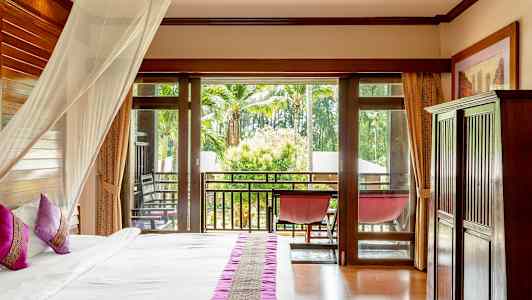 Ett hotellrum med en säng med rosa kuddar och en myggnätmantel, glasdörrar leder till en balkong med utsikt över palmer och grönska. (AI-genererad text)