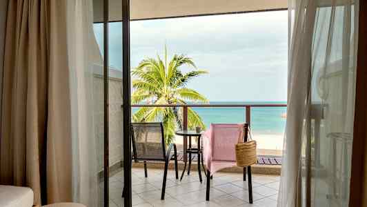 Utsikt från hotellrum mot en balkong med två stolar och ett bord, samt en strand och hav i bakgrunden. (AI-genererad text)