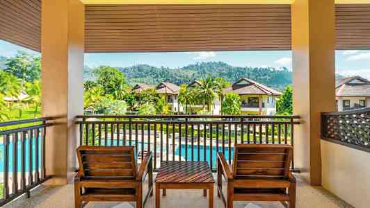 En uteplats med två trästolar och ett bord med utsikt över en grön trädgård, en pool och flerbostadshus. (AI-genererad text)