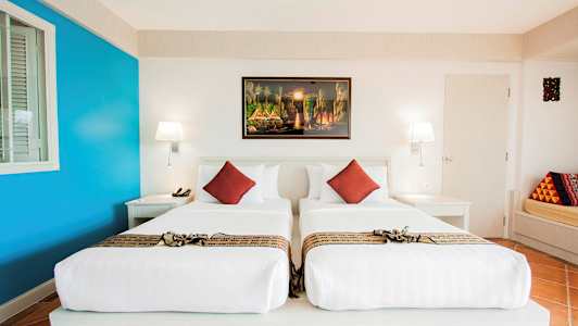 Ett hotellrum med två enkelsängar, vita sängkläder och dekorativa röda kuddar. På väggen hänger en färgglad tavla. (AI-genererad text)