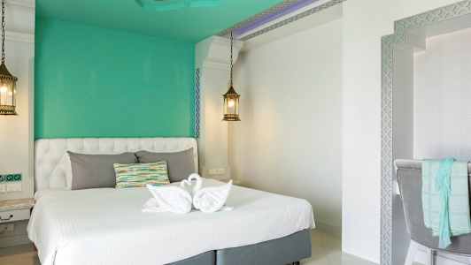 Ett elegant hotellrum med en vit dubbelsäng och gröna väggar. På sängen finns handdukar vikta till svanform och en färgglad kudde. Två hängande lampor skapar en behaglig atmosfär. (AI-genererad text)