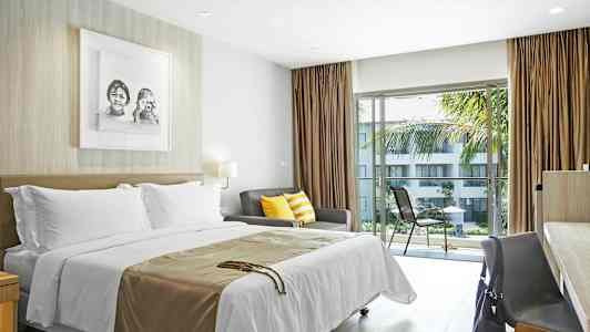 Ett hotellrum med säng, soffa med färgglada kuddar, och bord med stol vid fönster som vetter mot en balkong med utsikt över en trädgård. (AI-genererad text)