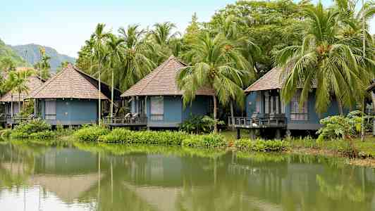Flera blå bungalower med halmtak ligger vid en grönskande sjö omgivna av palmer. (AI-genererad text)