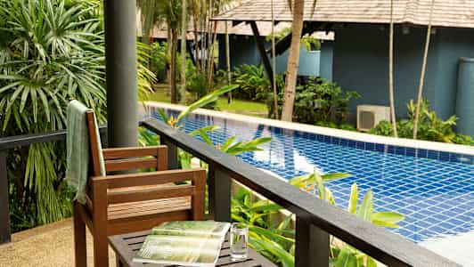 Utsikt från en veranda mot en pool omgiven av gröna växter och träd. (AI-genererad text)