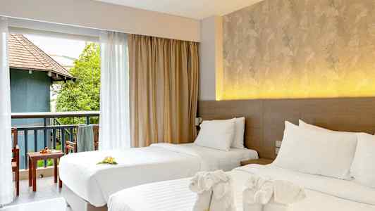 Två bäddade enkelsängar med prydnadshanddukar och blommor. Utsikt mot en möblerad balkong med grönska och byggnad. (AI-genererad text)