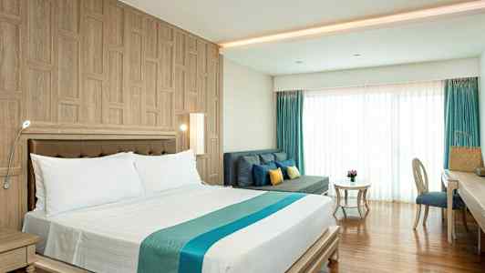 Ett hotellrum med en dubbelsäng, en soffa med kuddar, ett bord och en skrivbordsstol. Rummet har träpaneler på väggen och golv, samt stora fönster med gardiner. (AI-genererad text)