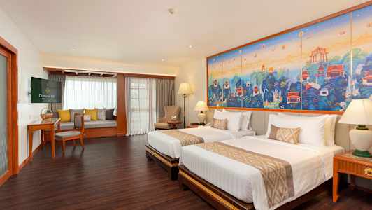 Ett hotellrum med två sängar, ett stort färgglatt konstverk på väggen och ett sittområde med fönster. (AI-genererad text)