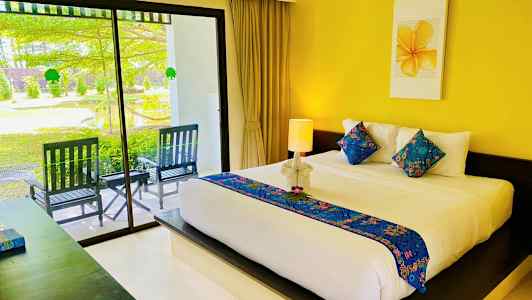 Ett hotellrum med en stor säng dekorerad med färgglada kuddar och ett ljust överkast. Sängen är placerad framför en vägg med en gul tavla ovanför. En glasdörr leder till en uteplats med trädgårdsmiljö. (AI-genererad text)