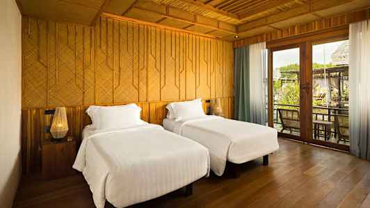 Två sängar med vita sängkläder ståendes i ett rum med trädetaljer och stora glasdörrar. (AI-genererad text)