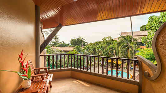 Bild av en balkong med ett träbord, stol och utsikt över en trädgård med pool. (AI-genererad text)
