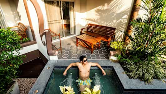 Person sitter i liten utomhuspool framför terrass med trämöbler och växter. (AI-genererad text)