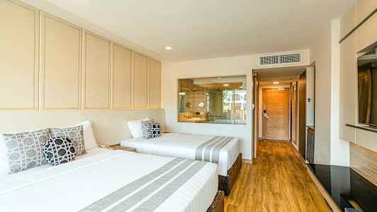 Ett hotellrum med två breda sängar bäddade med vita lakan och prydnadskuddar. Golvet är av trä och i bakgrunden finns en glasvägg med utsikt mot badrummet. (AI-genererad text)