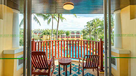 Utsikt från en balkong med två trästolar, ett litet bord och en vy över en pool omgiven av palmer och byggnader. (AI-genererad text)