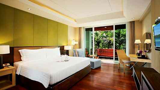 Sovrum med en stor säng, sittgrupp och skrivbord, samt utgång till en balkong som har utsikt över en trädgård. (AI-genererad text)