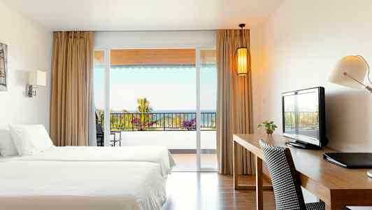 Ett hotellrum med två sängar, skrivbord, och utsikt mot en balkong med hav i bakgrunden. (AI-genererad text)