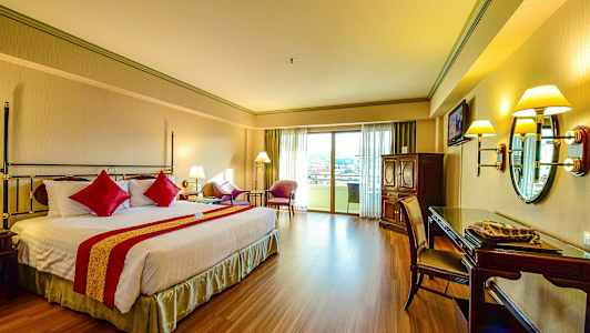 Ett hotellrum med en dubbelsäng och röd dekor, sittgrupp vid balkongdörren, och skrivbord med spegel. (AI-genererad text)