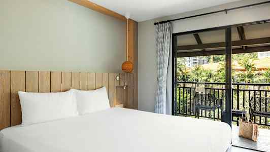 Bild på ett hotellrum med möbler i trä och en säng med vita lakan. En balkong med utsikt finns utanför. (AI-genererad text)