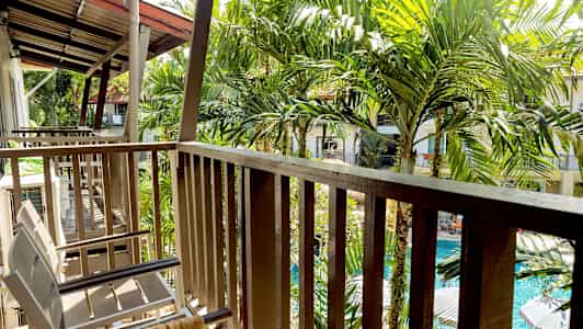 En balkong med stolar och räcke, omgiven av gröna palmer och med utsikt över en pool. (AI-genererad text)