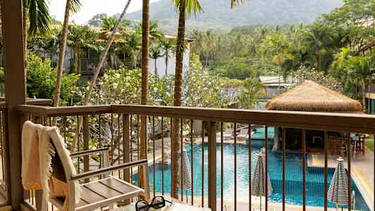 Utsikt från en balkong över en pool omgiven av palmer och gröna växter, med en stol och ett bord i förgrunden. (AI-genererad text)