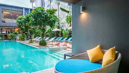 Modern pool med solstolar och växtlighet. En sittplats med kuddar i förgrunden. (AI-genererad text)