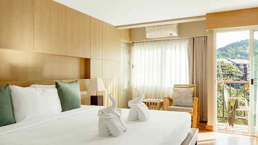 Ett välupplyst hotellrum med en stor säng täckt av vita lakan och kuddar. På sängen finns två handdukar vikta till formen av svanar. Rummet har en träinredning, en sittgrupp med kuddar och en balkong med utsikt mot grönska och byggnader. (AI-genererad text)