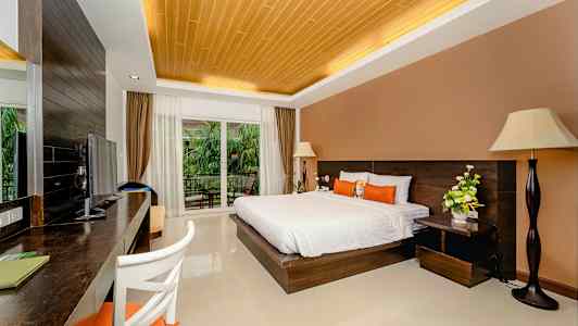 Ett hotellrum med en dubbelsäng, skrivbord, TV och stora glasdörrar med utsikt mot en balkong och grönska. (AI-genererad text)