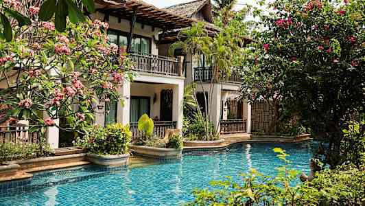 En pool med blått vatten omgiven av tropiska växter och en byggnad med balkonger. (AI-genererad text)