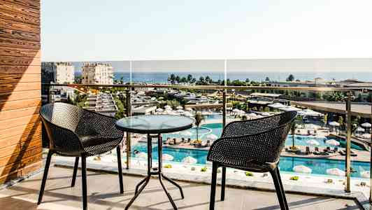 Balkong med två stolar och ett glasbord, utsikt över poolområde och hav. (AI-genererad text)