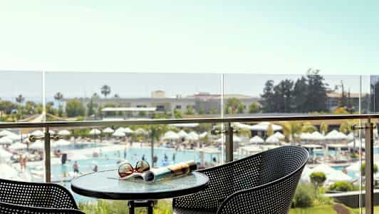 Utsikt från en balkong mot en poolområde med parasoller, omgiven av grönska. (AI-genererad text)