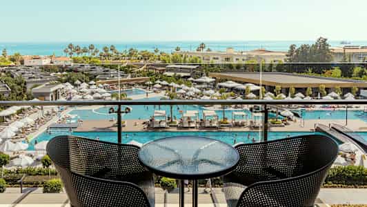 Vy från en balkong med ett bord och två stolar, utsikt över en pool och havet i bakgrunden. (AI-genererad text)