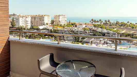 Utsikt från en balkong med bord och stolar över en pool och byggnader mot havet. (AI-genererad text)