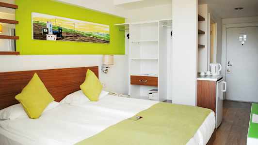 Hotellrum med en dubbelsäng, färgade kuddar, gröna dekorationsdetaljer och en öppen garderob. (AI-genererad text)