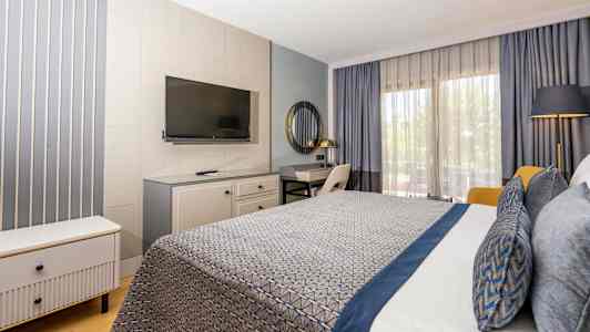 Hotellrum med modernt inredd interiör, inklusive säng med blå detaljer, skrivbord, TV och stora fönster med gardiner. (AI-genererad text)