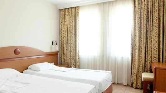 Ett hotellrum med två enkelsängar, en bord och en stol framför ett stort fönster med gardiner. (AI-genererad text)