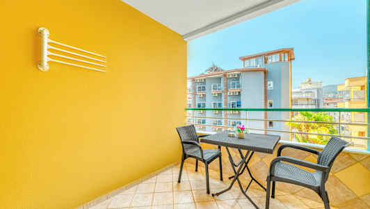 En balkong med ett gult vägg, ett bord med två stolar och en utsikt över andra byggnader. (AI-genererad text)