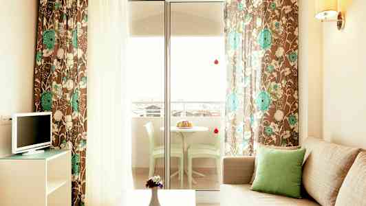 Soffa med grön kudde, tv och ett bord med vas framför. Blommiga gardiner och glasdörr till en terrass med bord och stolar. (AI-genererad text)