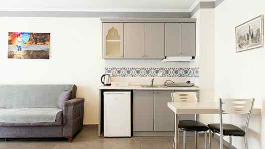 Ett rum med ett grått soffmöblemang, en kökshörna med vit kylskåp, gråa skåp och färgglada kakel. En matplats med bord och stolar står bredvid. (AI-genererad text)