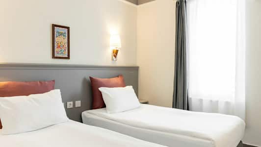 Två sängar med vita lakan och röda kuddar står i ett rum med ljus vägg och en vägglampa. Ett fönster med vita gardiner finns till höger. (AI-genererad text)