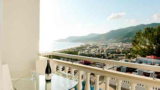 Balkong med glasbord, flaska vin och utsikt över stad och kust. (AI-genererad text)