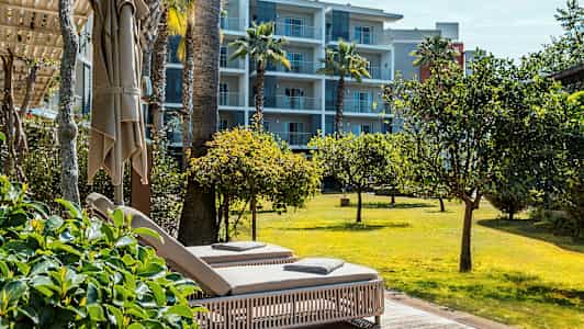 Två solstolar på en terrass med utsikt över en gräsmatta och träd, med ett lägenhetshotell i bakgrunden. (AI-genererad text)