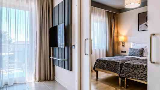 Modern hotellrum med två enkelsängar, en platt-TV på väggen och stora fönster med gardiner. (AI-genererad text)