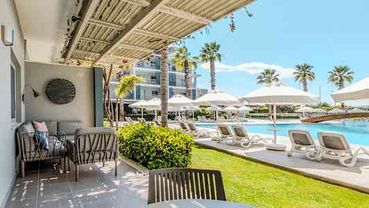 En utomhusveranda med sittmöbler och utsikt över en pool med solstolar och parasoller. (AI-genererad text)