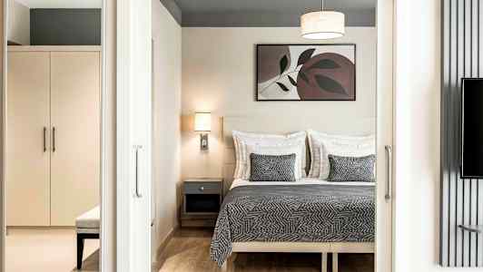 Ett sovrum med en dubbelbädd med grå quilt och matchande kuddar, en vägglampa, och en tavla ovanför sängen. Ett skåp och bänk syns i förgrunden. (AI-genererad text)