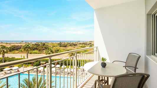 Utsikt från en balkong med stolar och bord, över ett poolområde med solstolar och palmer, samt vidare mot ett öppet landskap och havet. (AI-genererad text)