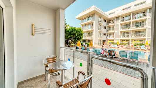 En balkong med bord och stolar vetter mot en pool och ett hotell. (AI-genererad text)