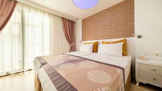 Hotellrum med en dubbelsäng, dekorerad med mönstrat överkast och kuddar. Fönster med genomskinliga gardiner och lila draperier i bakgrunden. (AI-genererad text)