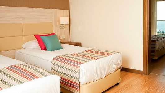 Två enkelsängar med randiga överkast och färgglada kuddar, i ett rum med trägolv och ljusa väggar. (AI-genererad text)