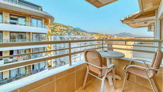 Balkong med två stolar och ett bord, med utsikt över byggnader och berg. (AI-genererad text)