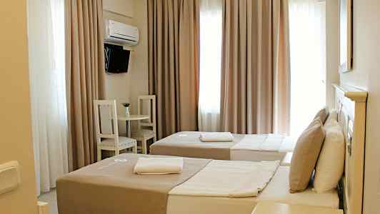 Ett hotellrum med två enkelsängar, beige sängkläder och kuddar. Ett litet bord med två stolar står vid väggen. Gardiner täcker fönstren, och på väggen finns en liten flatskärms-TV. (AI-genererad text)