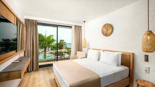 Ett hotellrum med en stor säng och en balkong med utsikt över en pool och palmträd. (AI-genererad text)